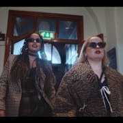A young Black woman and a young white woman in stylish clothes and sunglasses strutting down a hall