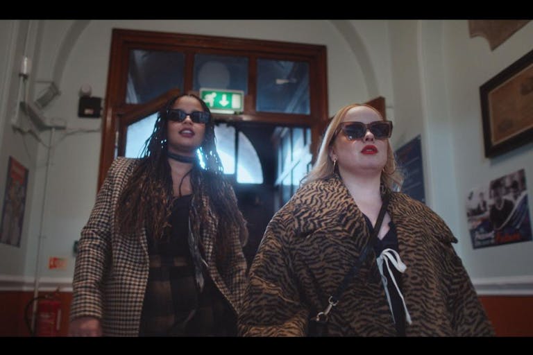 A young Black woman and a young white woman in stylish clothes and sunglasses strutting down a hall