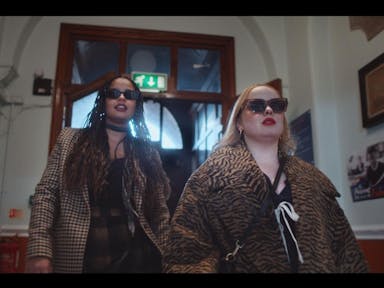 A young Black woman and a young white woman in stylish clothes and sunglasses strutting down a hall