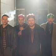 Four white men swaggering down an alley in cheap suits and ties, smoking, some wearing Christmas cracker party hats