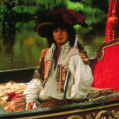 A white man in 17th Century regal, flamboyant attire, sitting in a plush lake boat