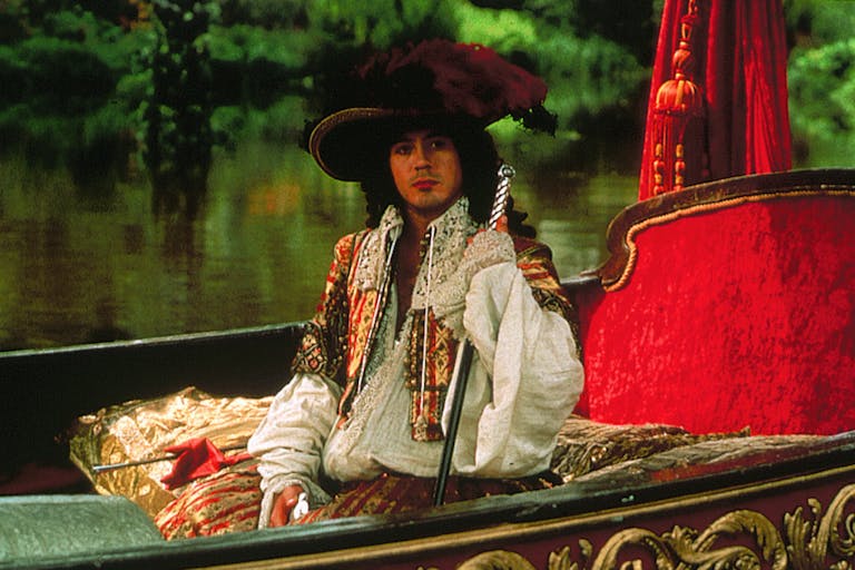 A white man in 17th Century regal, flamboyant attire, sitting in a plush lake boat