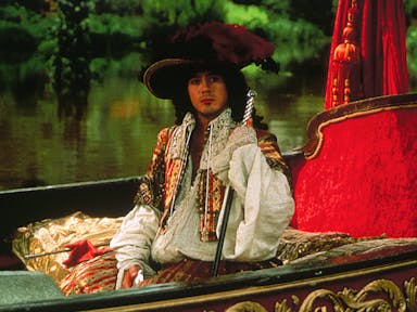 A white man in 17th Century regal, flamboyant attire, sitting in a plush lake boat