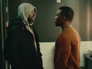 Two Black men standing facing each other looking serious, one wearing a grey hoodie and black coat, the other wearing a burnt orange sweater