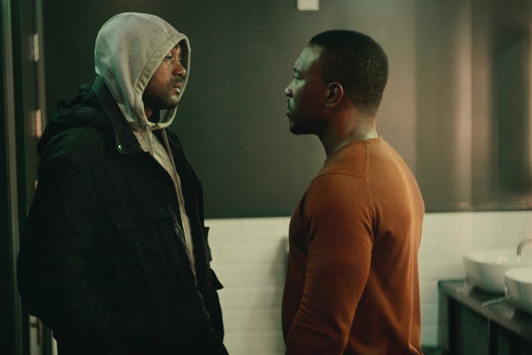 Two Black men standing facing each other looking serious, one wearing a grey hoodie and black coat, the other wearing a burnt orange sweater