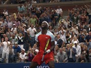 A Black male tennis player in red sports shirt and shorts holding a racket celebrating with lots of energy as a numerous rows of spectators celebrate behind him