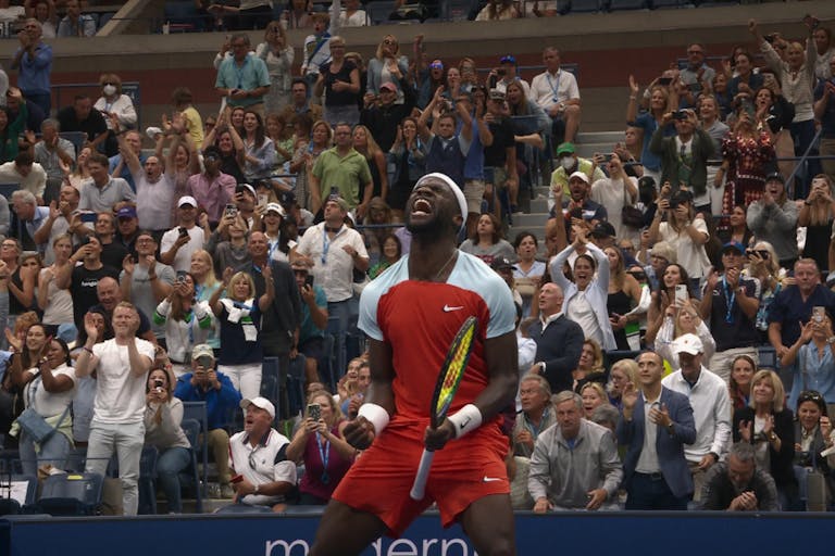 A Black male tennis player in red sports shirt and shorts holding a racket celebrating with lots of energy as a numerous rows of spectators celebrate behind him