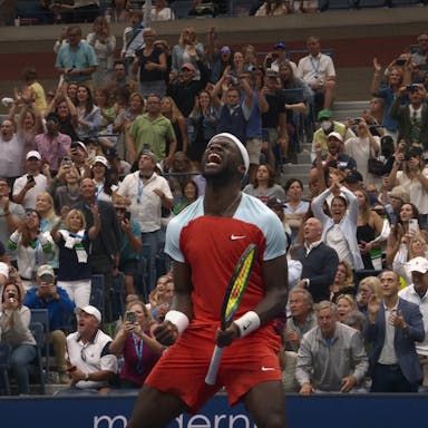 A Black male tennis player in red sports shirt and shorts holding a racket celebrating with lots of energy as a numerous rows of spectators celebrate behind him