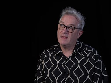 A middle aged white man with patterned shirt, black glasses and quiffed grey hair
