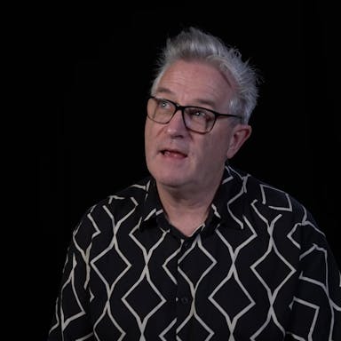 A middle aged white man with patterned shirt, black glasses and quiffed grey hair