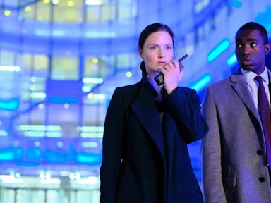 Woman speaks into a walkie-talkie next to a man in a suit, both against a background lit up by blue lights