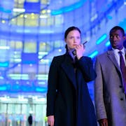 Woman speaks into a walkie-talkie next to a man in a suit, both against a background lit up by blue lights