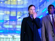 Woman speaks into a walkie-talkie next to a man in a suit, both against a background lit up by blue lights