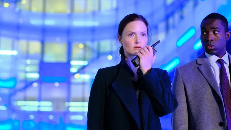 Woman speaks into a walkie-talkie next to a man in a suit, both against a background lit up by blue lights