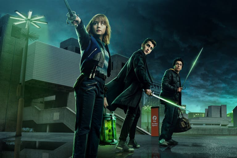 Three young adults stand against an industrial night, green-tinged sky, holding swords.