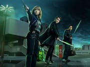 Three young adults stand against an industrial night, green-tinged sky, holding swords.