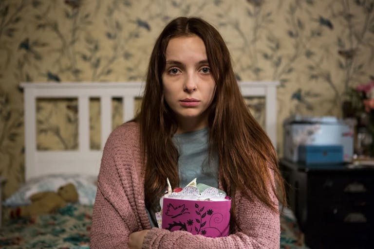 A tired-looking young white woman sitting on a bed clutching a stuffed diary