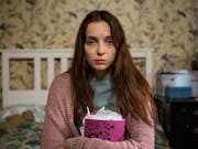 A tired-looking young white woman sitting on a bed clutching a stuffed diary