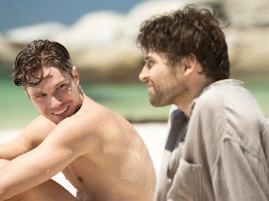 Topless young man looks at another young man, wearing a grey shirt, and smiles