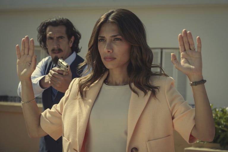 A man stands behind a woman with his hands up, pointing a gun at her back