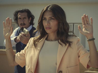 A man stands behind a woman with his hands up, pointing a gun at her back 