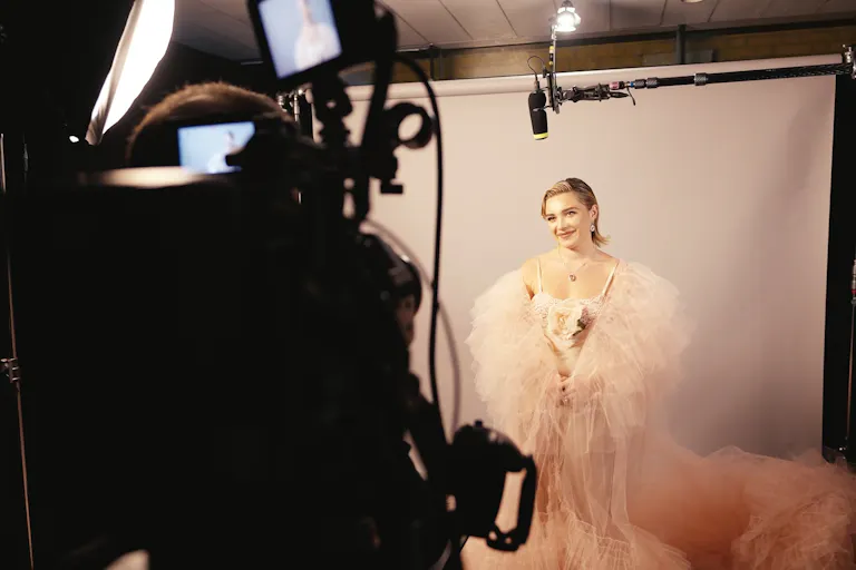 Glamourous young woman wearing a light pink dress and tulle cape stands in front of a camera