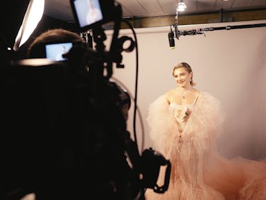 Glamourous young woman wearing a light pink dress and tulle cape stands in front of a camera