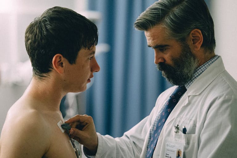 A middle aged white man in a white doctors coat applying wires and monitors to bare torso of a young white man