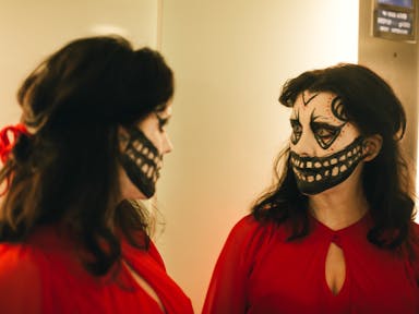A white woman with brown hair wearing a bright red dress, looks in the mirror with her face painted in a creepy, black and white mask style, with a large mouth extending across her face exposing teeth