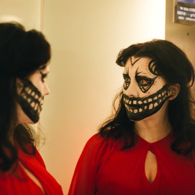 A white woman with brown hair wearing a bright red dress, looks in the mirror with her face painted in a creepy, black and white mask style, with a large mouth extending across her face exposing teeth