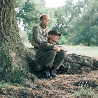 Two middle aged white men sitting on large tree roots, wearing muted outdoor clothes