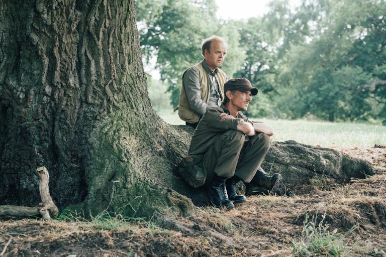Two middle aged white men sitting on large tree roots, wearing muted outdoor clothes
