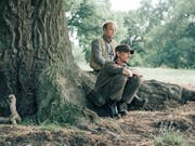 Two middle aged white men sitting on large tree roots, wearing muted outdoor clothes