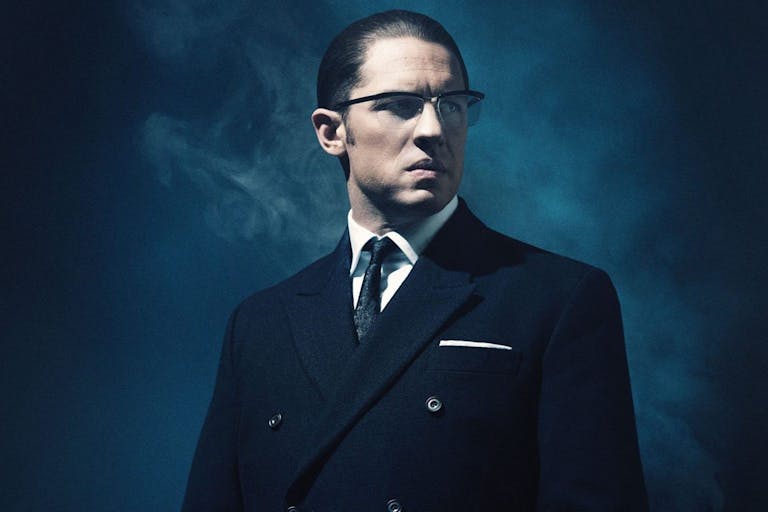 A white man with slicked back dark hair wearing glasses and a sharp dark suit, looking menacing