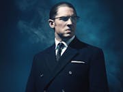 A white man with slicked back dark hair wearing glasses and a sharp dark suit, looking menacing