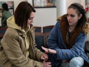 Two teenage girls in grungy clothing sit next to each other talking in a doctor's waiting room.