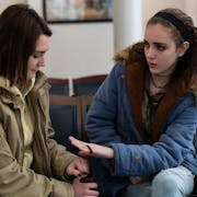 Two teenage girls in grungy clothing sit next to each other talking in a doctor's waiting room.
