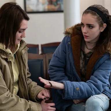 Two teenage girls in grungy clothing sit next to each other talking in a doctor's waiting room. 