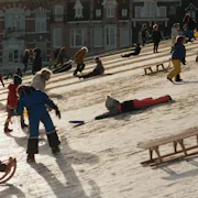 Young children playing on a snowy slope in snow suits and using wooden sledges