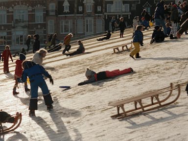 Young children playing on a snowy slope in snow suits and using wooden sledges