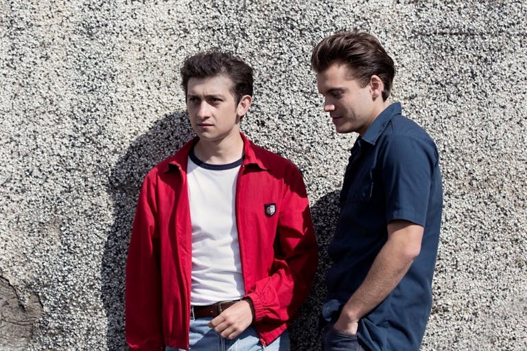 Two white teen boys with slicked back hair lean again a pebble dash wall