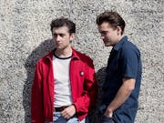 Two white teen boys with slicked back hair lean again a pebble dash wall