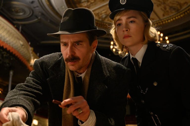 Man with a moustache and 1950s style hat stands next to a blonde woman in a police uniform, they look like they are inside an empty theatre. 