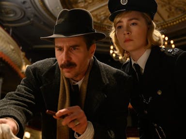 Man with a moustache and 1950s style hat stands next to a blonde woman in a police uniform, they look like they are inside an empty theatre. 