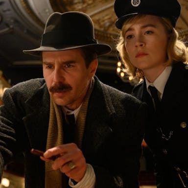 Man with a moustache and 1950s style hat stands next to a blonde woman in a police uniform, they look like they are inside an empty theatre. 