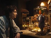 A young white man stares at a tiny bright orange man with green hair trapped under a glass dome on a desk