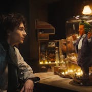 A young white man stares at a tiny bright orange man with green hair trapped under a glass dome on a desk