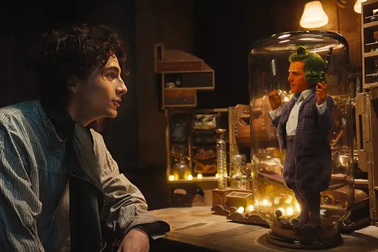 A young white man stares at a tiny bright orange man with green hair trapped under a glass dome on a desk