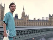 A young skinny white man in hospital scrubs stands on an empty bridge in London with the Parliament building behind him