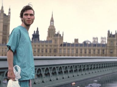 A young skinny white man in hospital scrubs stands on an empty bridge in London with the Parliament building behind him 